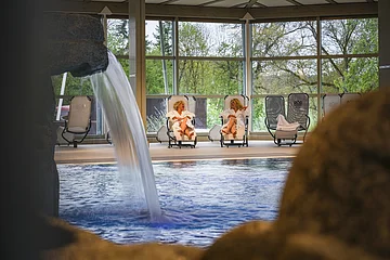 Zwei Gäste im Bademantel liegen gemütlich auf den Ruheliegen, die um das Schwimmbecken verteilt sind.