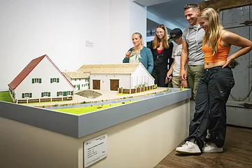 Vier Gäste in der Abteilung Möbel aus dem 17. Jahrhundert. Sie stehen auf der rechten Seite und blicken auf das Modell eines Jura-Bauernhofes.