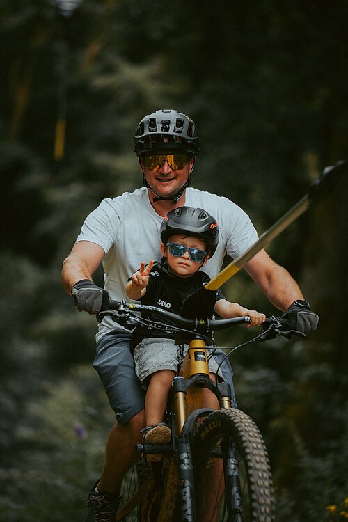 Auf dem Bild ist ein Vater mit seinem Sohn auf einem Fahrrad zu sehen, welche mithilfe eines Liftes hochgezogen werden.