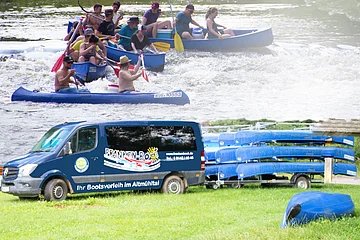 Eine Gruppe von Menschen paddelt in Kanus auf einem Fluss. Darunter steht ein Transporter mit Kanus auf einem Anhänger.