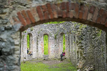 Zwei Personen sitzen in den Fensternischen einer alten Steinruine mit grüner Vegetation im Hintergrund.