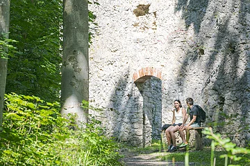 Zwei Personen sitzen auf einer Bank vor einer alten Steinruine im Wald.