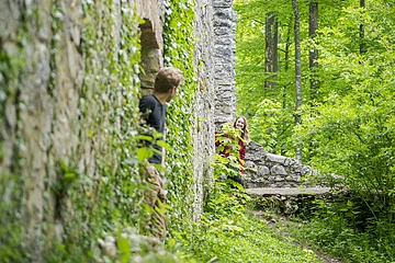 Zwei Personen schauen um eine mit Efeu bewachsene Steinmauer in einem Waldgebiet.