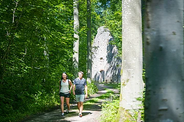 Zwei Personen wandern auf einem Waldweg, im Hintergrund eine alte Steinruine.