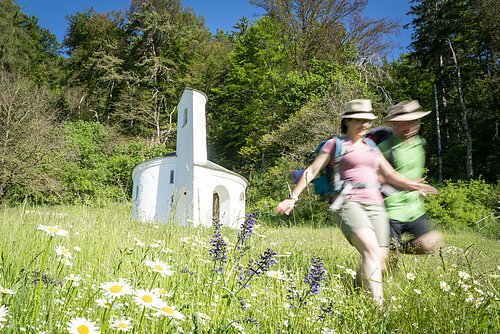 St. Gunthildiskapelle mit Wanderpärchen Zwei Personen wandern auf einer Wiese mit Blumen vor einer kleinen weißen Kapelle im Wald.