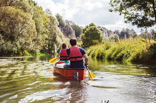 Zwei Personen mit roten Schwimmwesten paddeln in einem roten Kanu auf einem Fluss, umgeben von grüner Natur.