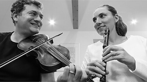 Auf dem Bild ist eine Frau und ein Mann zu sehen. Der Mann hat eine Geige, während die Frau eine Klarinette hält.