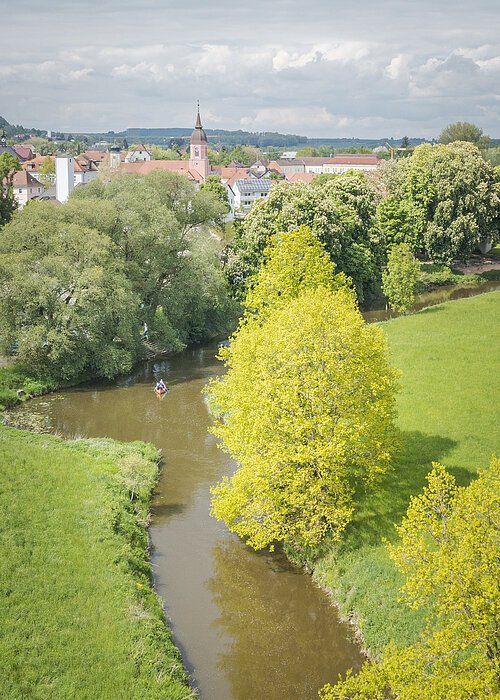 Fluss mit grünen Bäumen und Wiesen, im Hintergrund Häuser und Kirchturm unter bewölktem Himmel