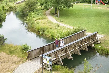 Drohnenaufnahme von Möhren. Im Vordergrund sind 2 Wanderer auf einer Holzbrücke über den Möhrenbach zu sehen. Im Hintergrund thront Schloss Möhren über dem Dorf.
