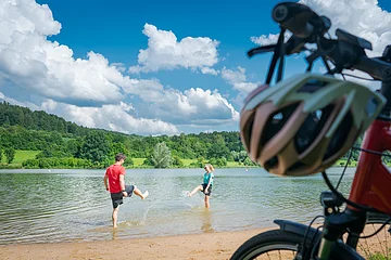 Im Vordergrund stehen rechts zwei Fahrräder, dahinter liegt ein kurzer Strand an einem Seeufer. Im seichten Wasser stehen ein Mann und in eine Frau in kurzen Hosen und T-Shirts, die versuchen, sich gegenseitig nass zu spritzen. Am Ufer gegenüber liegen eine Wiese und Wald.