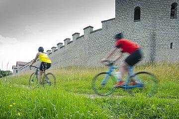Zwei Gravelbiker vorne mit gelben Shirt, dahinter mit rotem Shirt fahren seitlich am Kastell entlang. Der Himmel ist grau bedeckt, die Wiesen saftig grün.