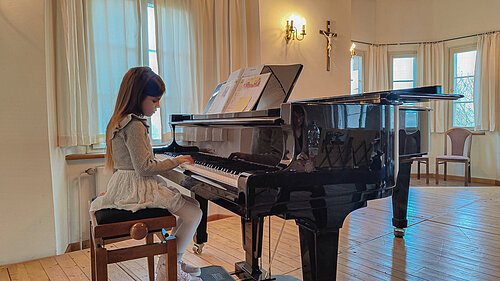 Junges Mädchen sitzt an einem Flügel und spielt darauf. Der Flügel steht auf einer kleinen Bühne.