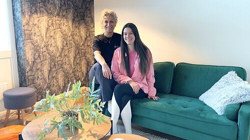 Bahar und Alex Zwei Yogalehrerinnen sitzen auf einem grünen Sofa. Im Vordergrund steht ein runder Tisch mit einer grünen Pflanze.