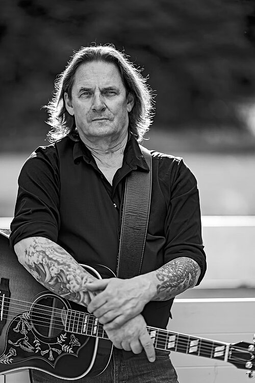 Auf dem Bild ist der Musiker Rusty Stone mit seiner Gitarre in der Natur zu sehen.